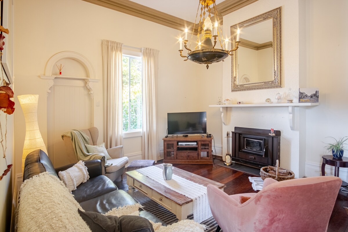 A cozy living area is presented with a leather couch and armchairs, complemented by a wooden coffee table. A flat-screen TV is mounted on a white wall, above which a mirror enhances the space. Soft light filters through sheer curtains, creating a warm ambiance.