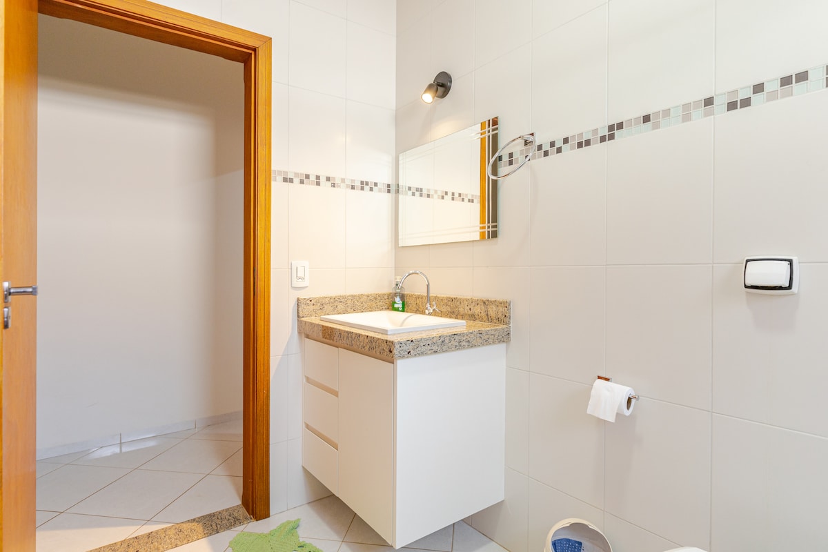 A spacious bathroom features a modern sink with a granite countertop and ample storage below. A well-lit mirror reflects the serene tile design, which includes a decorative border. The bathroom is equipped with a shower and essential toiletries arranged for convenience.