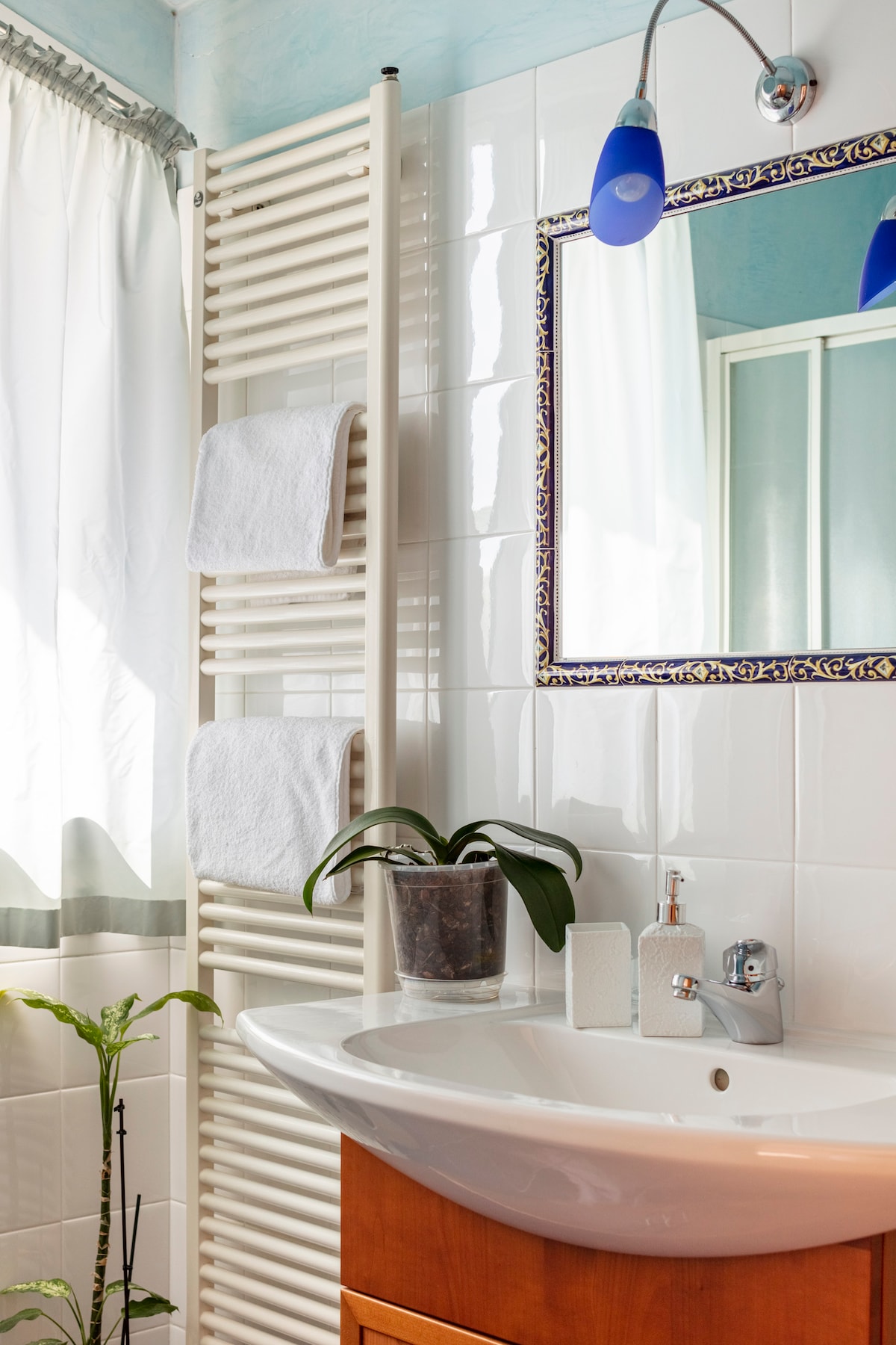 The bathroom features a white tiled wall with a mirror framed in decorative tile. A wooden vanity supports a rounded sink and modern faucet. Two white towels are neatly hung on a heated towel rack. A green plant adds a touch of nature beside the sink.