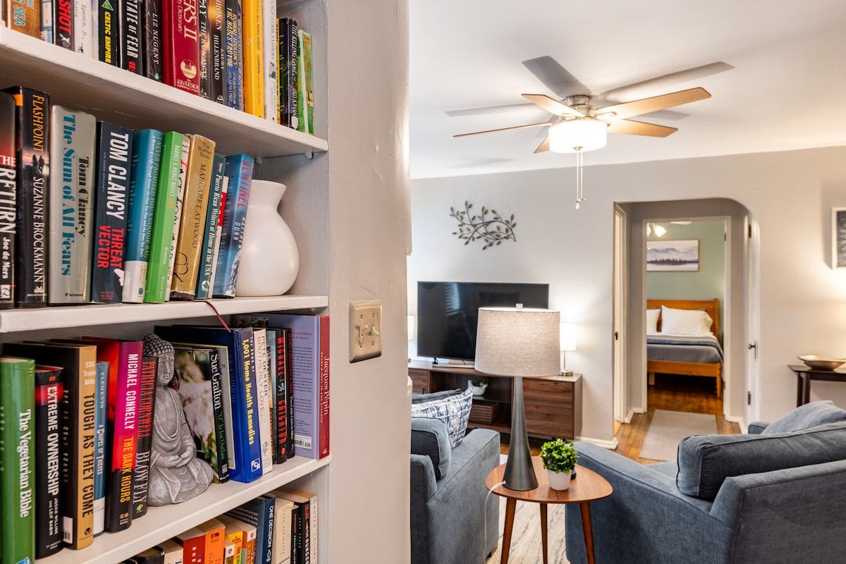 A living area features a soft blue couch paired with a wooden side table showcasing a small plant. A television stands on a modern console, while a ceiling fan provides airflow. A comfortable bedroom is visible in the background, contributing to the spacious ambiance.