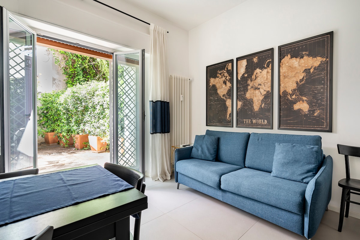 The living area features a comfortable blue sofa facing a black dining table. Two doors open to a private garden, creating a seamless connection between indoor and outdoor spaces. World maps are displayed on the walls, adding a touch of decor to the room.