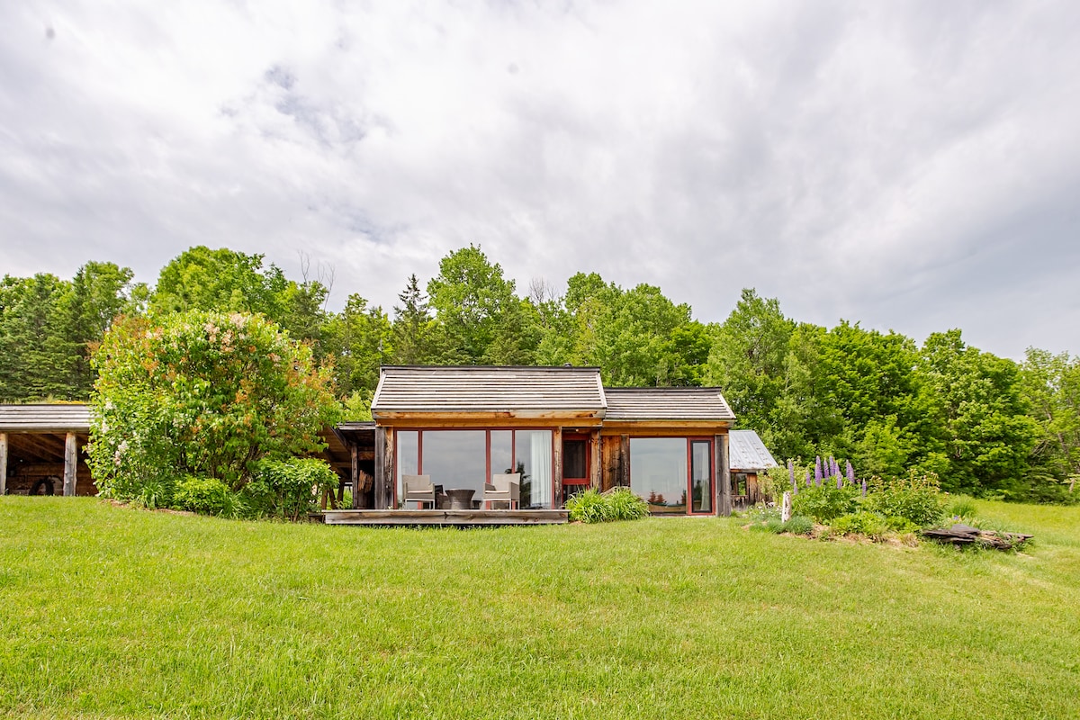 A charming home is set amidst lush greenery, featuring large windows that invite natural light. The exterior is complemented by a spacious deck with seating options, looking out over a grassy area. Vegetation borders the property, enhancing the serene environment.
