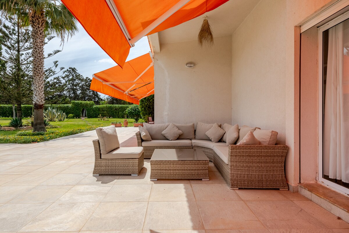 A spacious outdoor seating area is furnished with a comfortable sectional sofa and a glass coffee table, positioned on a tiled patio. Large, orange awnings provide shade, while lush greenery and garden elements are visible in the background.
