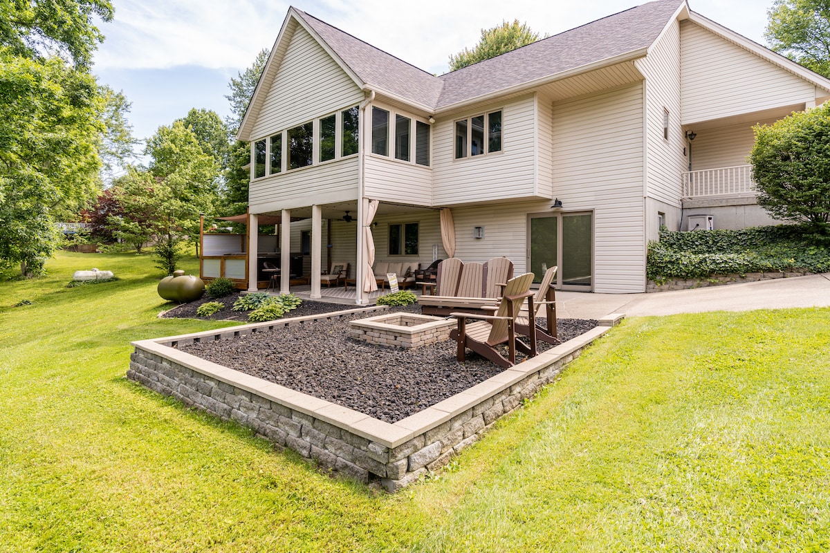 The exterior of the home features a manicured lawn with a gravel area and outdoor seating. Comfortable wooden chairs are arranged around a fire pit, surrounded by neatly maintained landscaping. Trees and greenery enhance the peaceful country setting of the property.