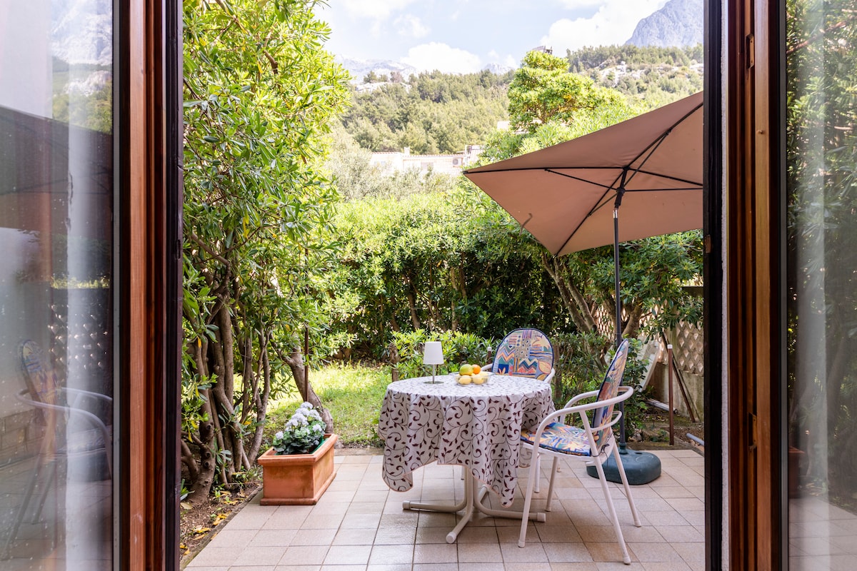 A private terrace is showcased, featuring a round table covered with a patterned tablecloth, surrounded by two chairs. An umbrella provides shade over the seating area, while greenery and a flower pot enhance the serene outdoor space. Views of distant mountains are visible in the background.