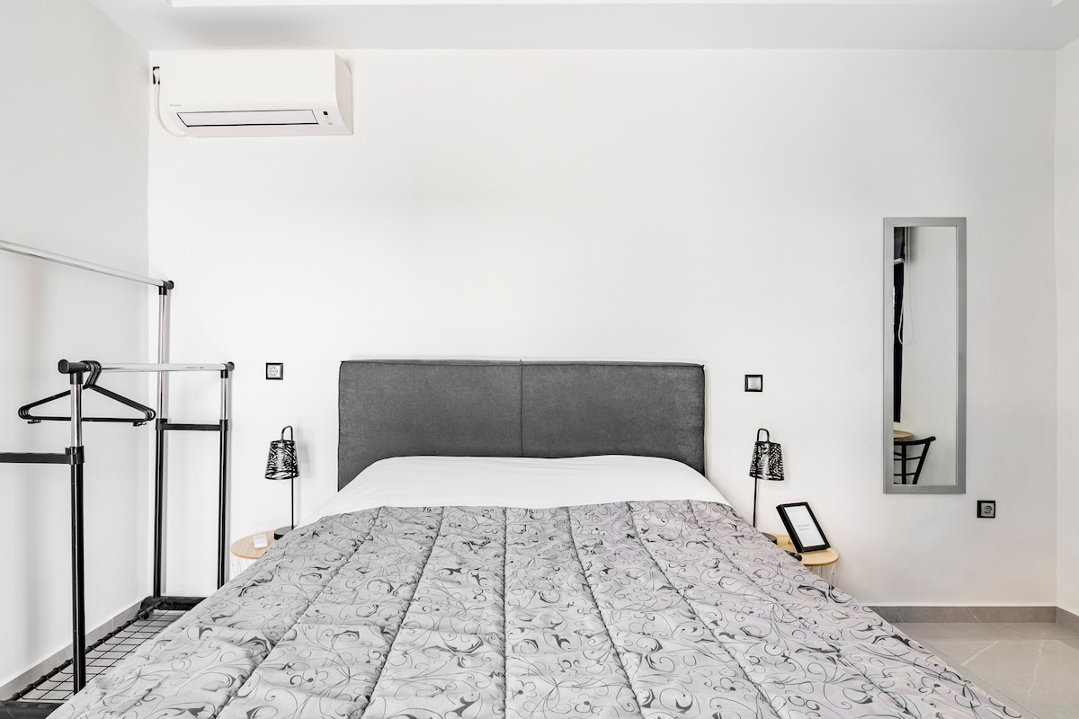 A modern sleeping area is centered around a spacious bed with a patterned gray quilt. A mirrored wall hangs nearby, reflecting the simple, stylish decor. Minimalist bedside tables with lamps and a clothing rack complete the uncluttered look of the room.