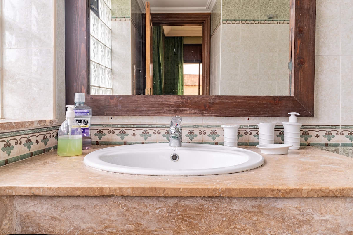 A marble countertop supports a circular sink with a chrome faucet, surrounded by an intricately designed backsplash. Nearby, various toiletries are organized, including a soap dispenser and a small bottle. A decorative mirror reflects the overall bathroom space.
