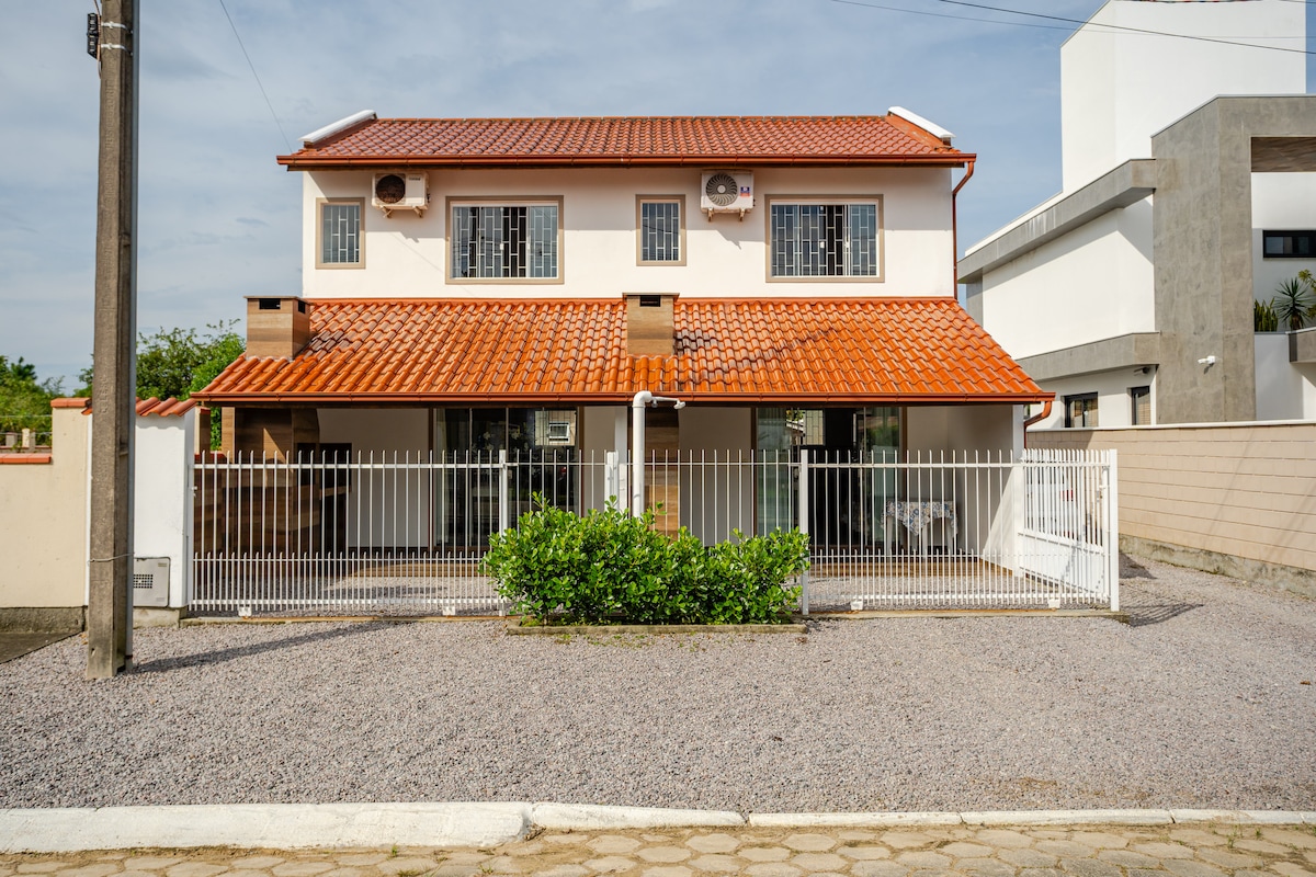 The two-story house features a red-tiled roof and a spacious front yard surrounded by a white fence. Large glass doors are visible, leading to a welcoming indoor space. Air conditioning units are seen on each upper window, ensuring comfort throughout the home.