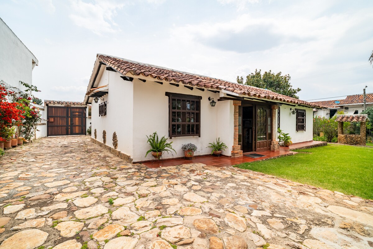 A charming colonial-style home features a spacious stone pathway leading to the entrance. The exterior showcases whitewashed walls and a traditional tiled roof. Lush greenery and flowering plants are visible, enhancing the inviting atmosphere. A private parking area is discernible on the left.