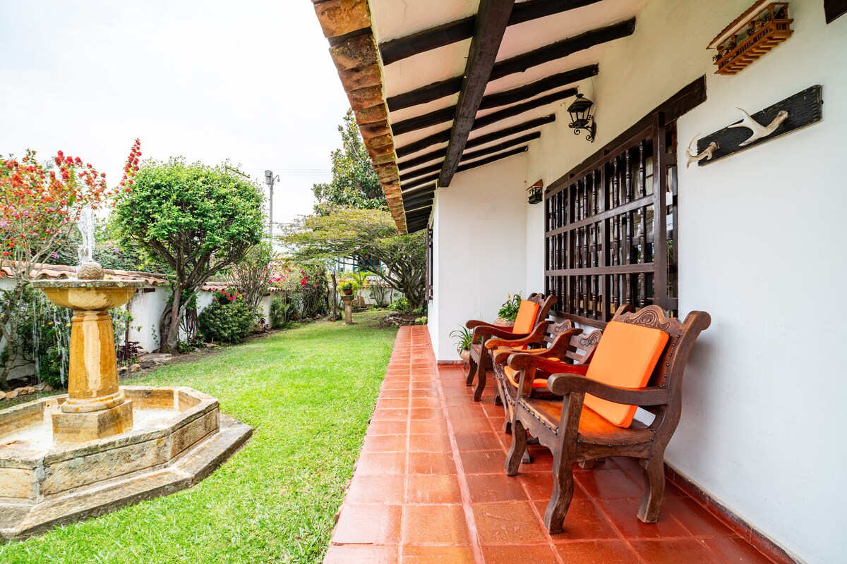 A peaceful patio area is set with two wooden chairs featuring vibrant orange cushions. A stone fountain adorned with greenery serves as a focal point in the adjacent yard, which is accented by colorful flowers and well-maintained grass.