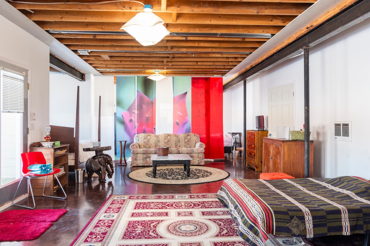 A spacious living area features exposed wooden beams and a vibrant rug, creating a bright environment. A comfortable sofa with floral patterns and a wooden cabinet are visible, along with a table and a chair. Colorful artwork adds interest to the background.