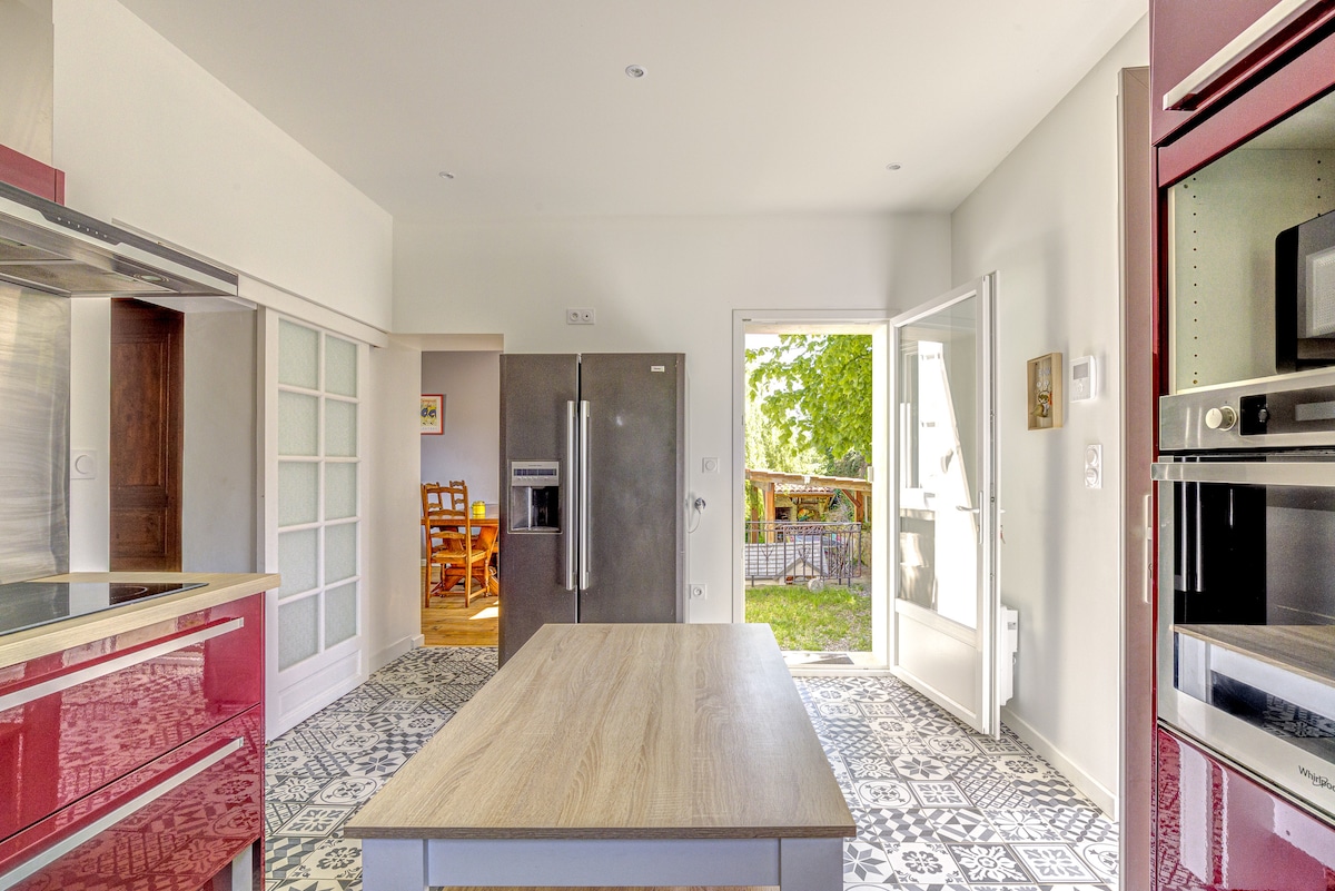A modern kitchen is shown with sleek red cabinetry and a large wooden island at its center. Natural light enters through the open door leading to the outside area. A glimpse of dining space is visible through the doorway, with patterned tile flooring throughout.