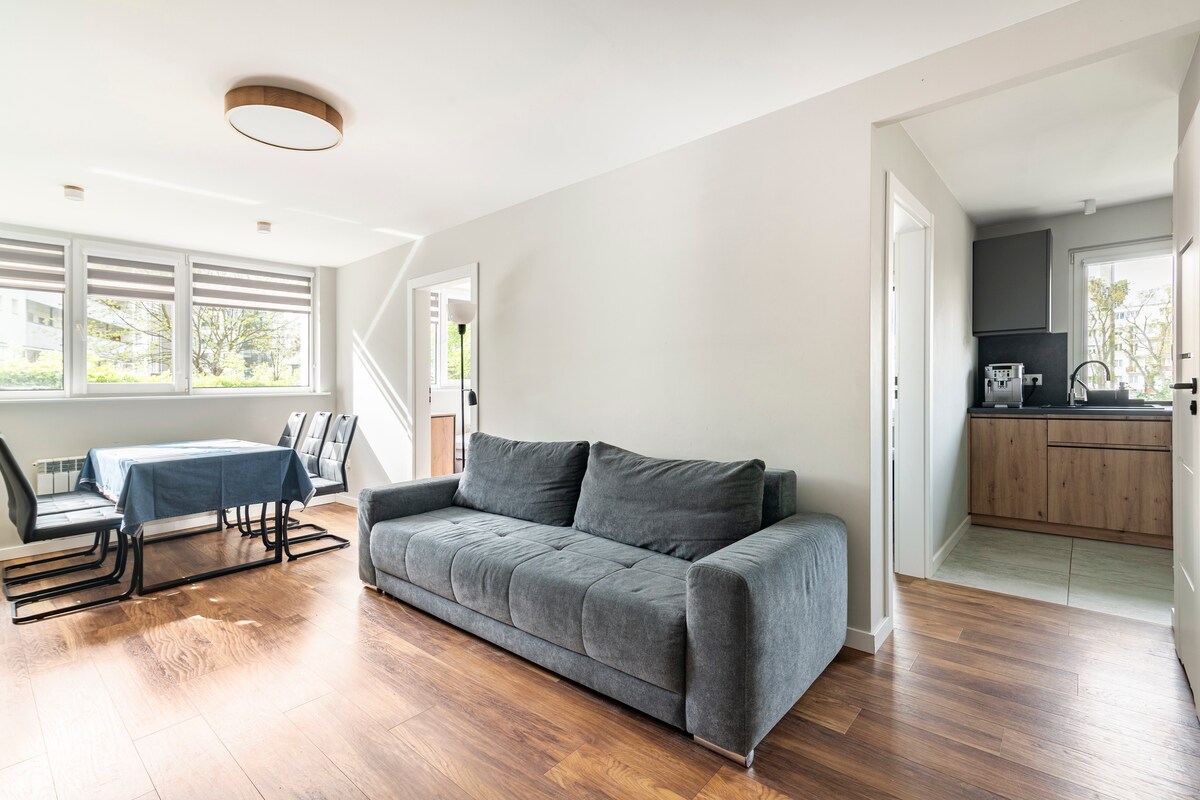 A spacious living area is presented with a dark gray sofa facing a light wood floor. A dining table set for six is visible in the background, with large windows allowing natural light to illuminate the room. A modern kitchen can be seen through an open doorway.
