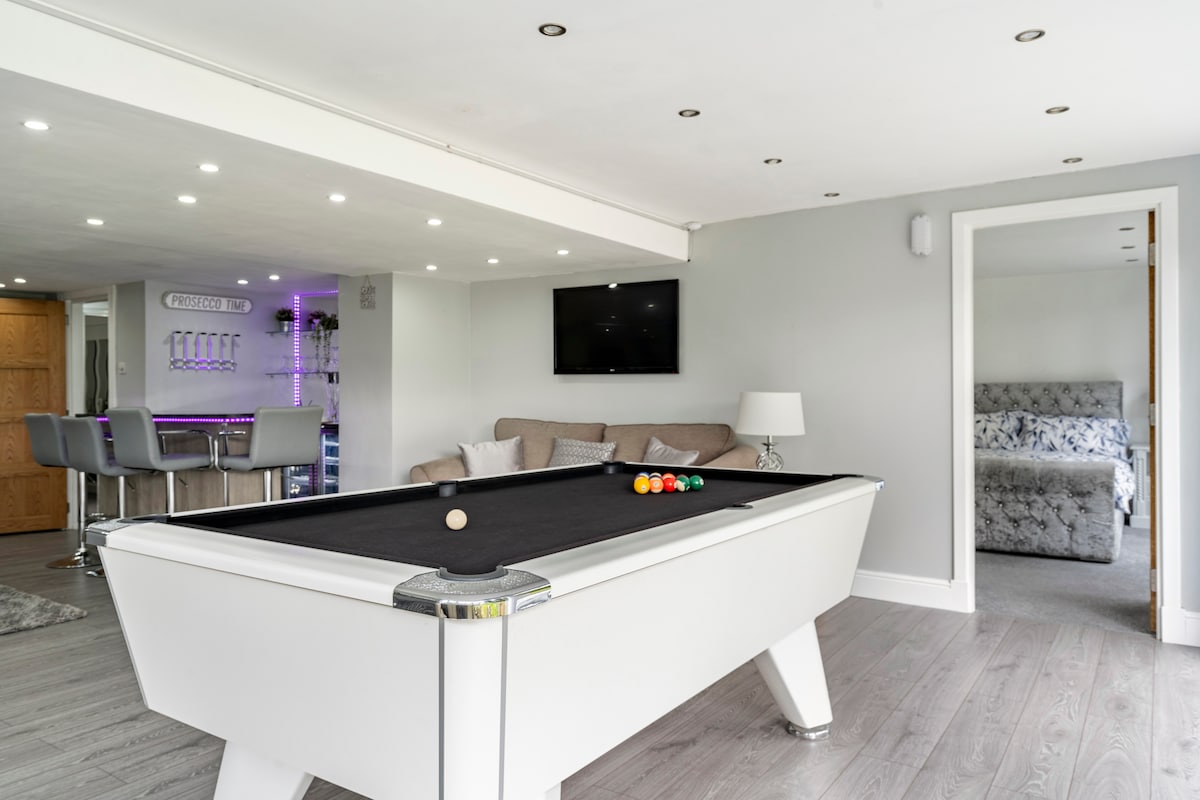 A modern pool table is positioned in a well-lit room, creating an inviting entertainment area. The space features a comfortable sectional sofa and a flat-screen television. Bright recessed lighting enhances the neutral-toned walls, while a stylish bar area can be seen in the background.
