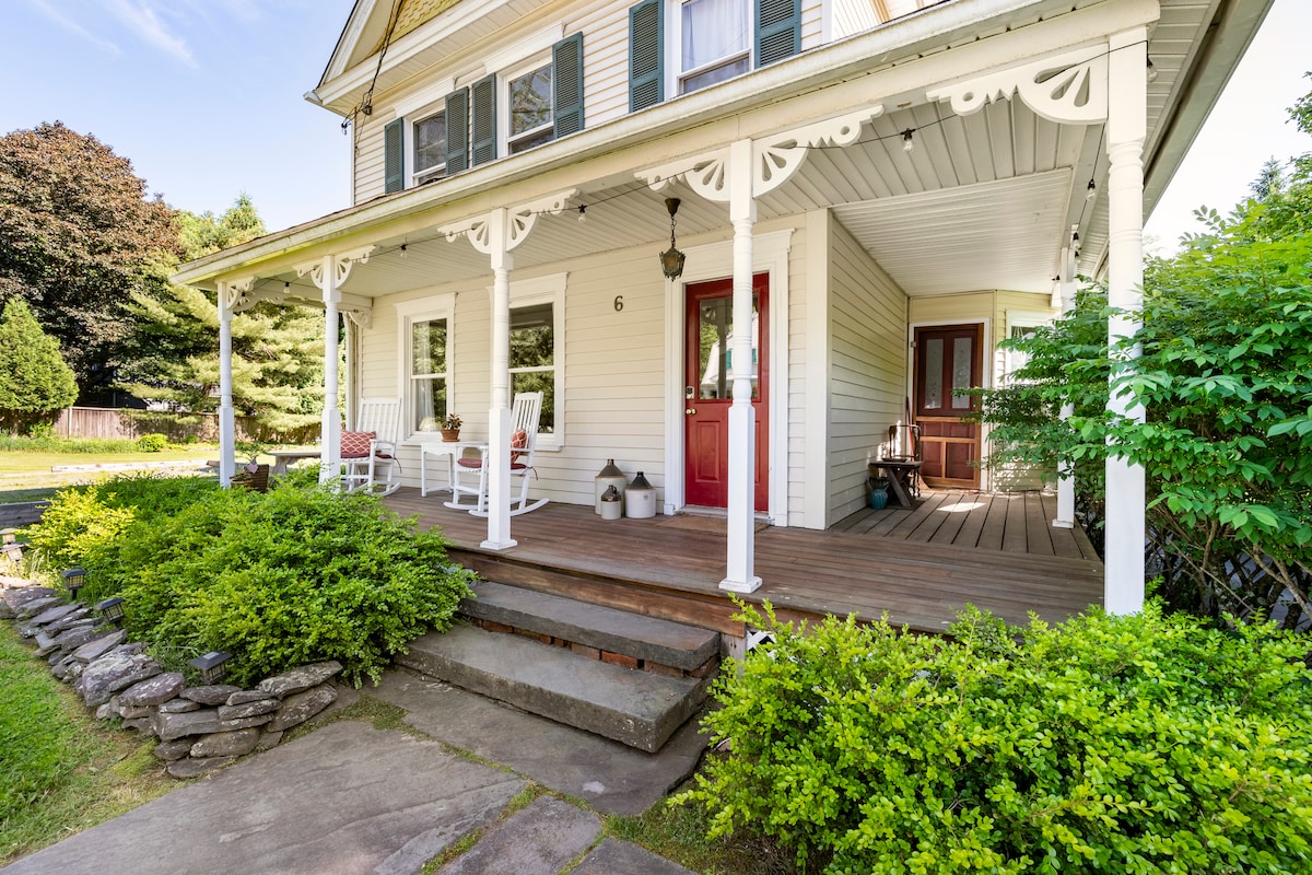 The charming front porch features multiple rocking chairs, framed by manicured greenery. Steps lead to a welcoming entrance, with decorative stone accents and potted plants adding to the inviting atmosphere. The home’s light-colored exterior and contrasting green shutters create a classic aesthetic.