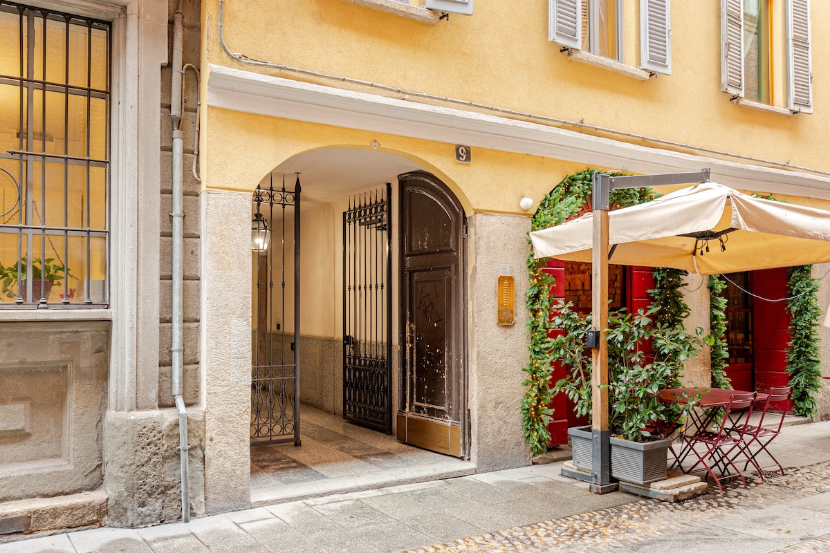 The entrance to the building is framed by charming yellow walls, with an ornate black gate leading into a well-maintained corridor. A small outdoor seating area is visible to the side, featuring a table with red chairs under a beige umbrella, providing a welcoming touch.