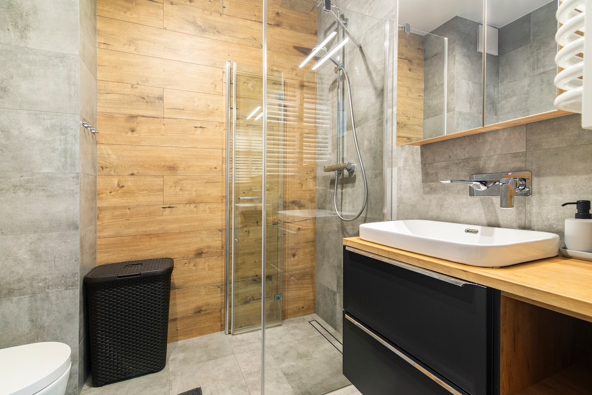 The modern bathroom features a walk-in shower enclosed by clear glass panels and equipped with a rain showerhead. A sleek black vanity with a white sink is positioned beside a large mirror. Distinctive wood accents on the walls complement the stone-like floor.