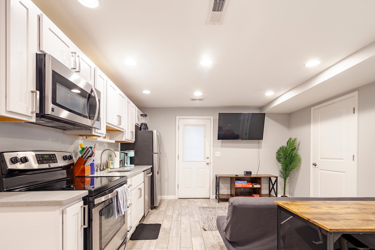 A well-equipped kitchen is visible, featuring white cabinetry and stainless steel appliances. The space includes a microwave, oven, and refrigerator, with a TV mounted on the wall and a door leading outside. Natural light fills the area, complemented by light grey tiled flooring.