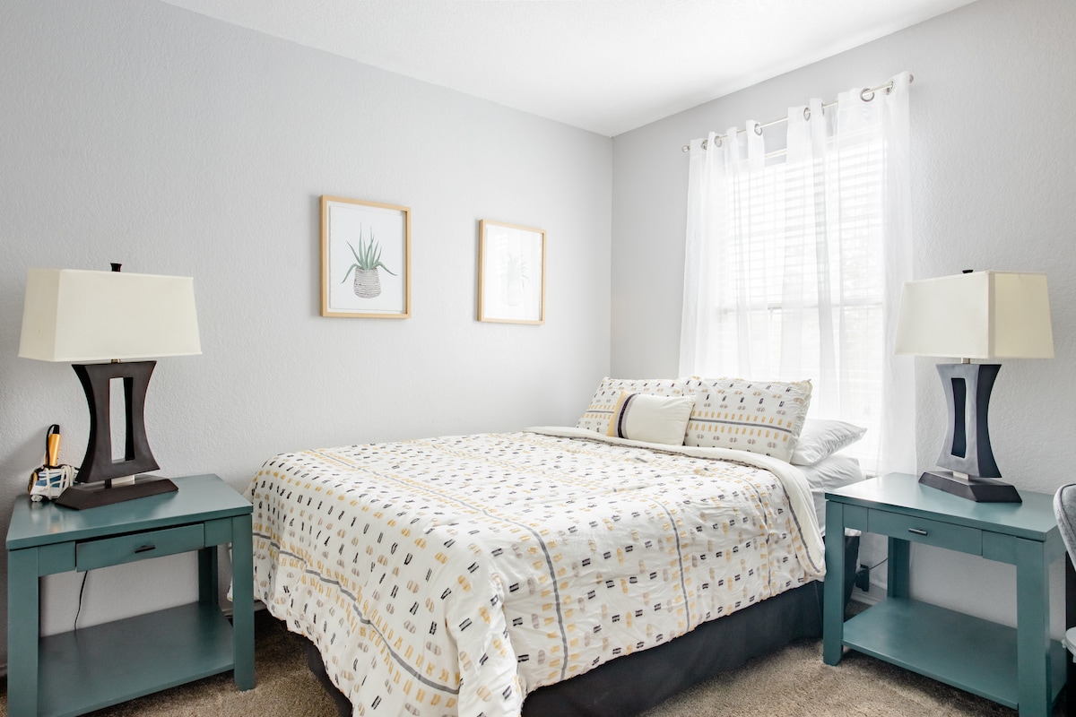 An inviting bedroom is furnished with a spacious bed dressed in a patterned duvet cover. Two bedside tables with lamps flank the bed, and light filters through sheer curtains. Simple artwork hangs on the pale walls, contributing to a calm atmosphere.