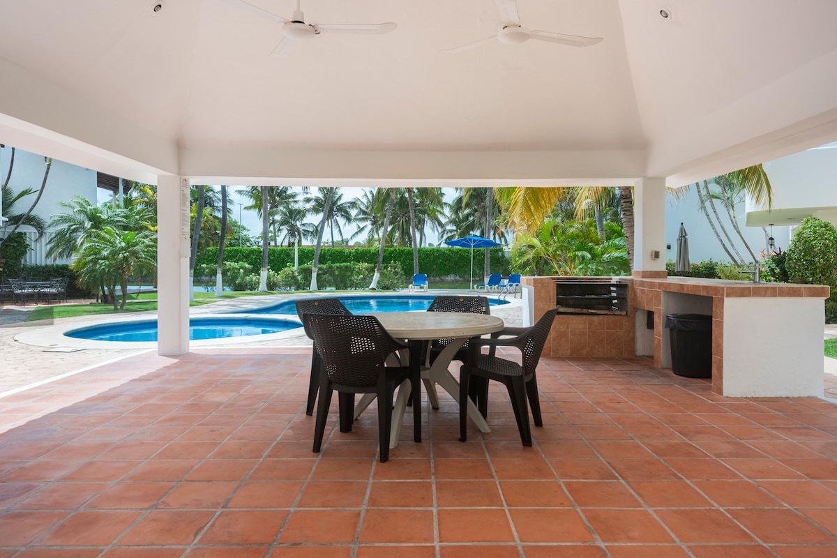 An outdoor seating area features a round table surrounded by six dark chairs, situated under a spacious canopy. A turquoise swimming pool is visible in the background, surrounded by palm trees and lounge chairs, creating a relaxed atmosphere.