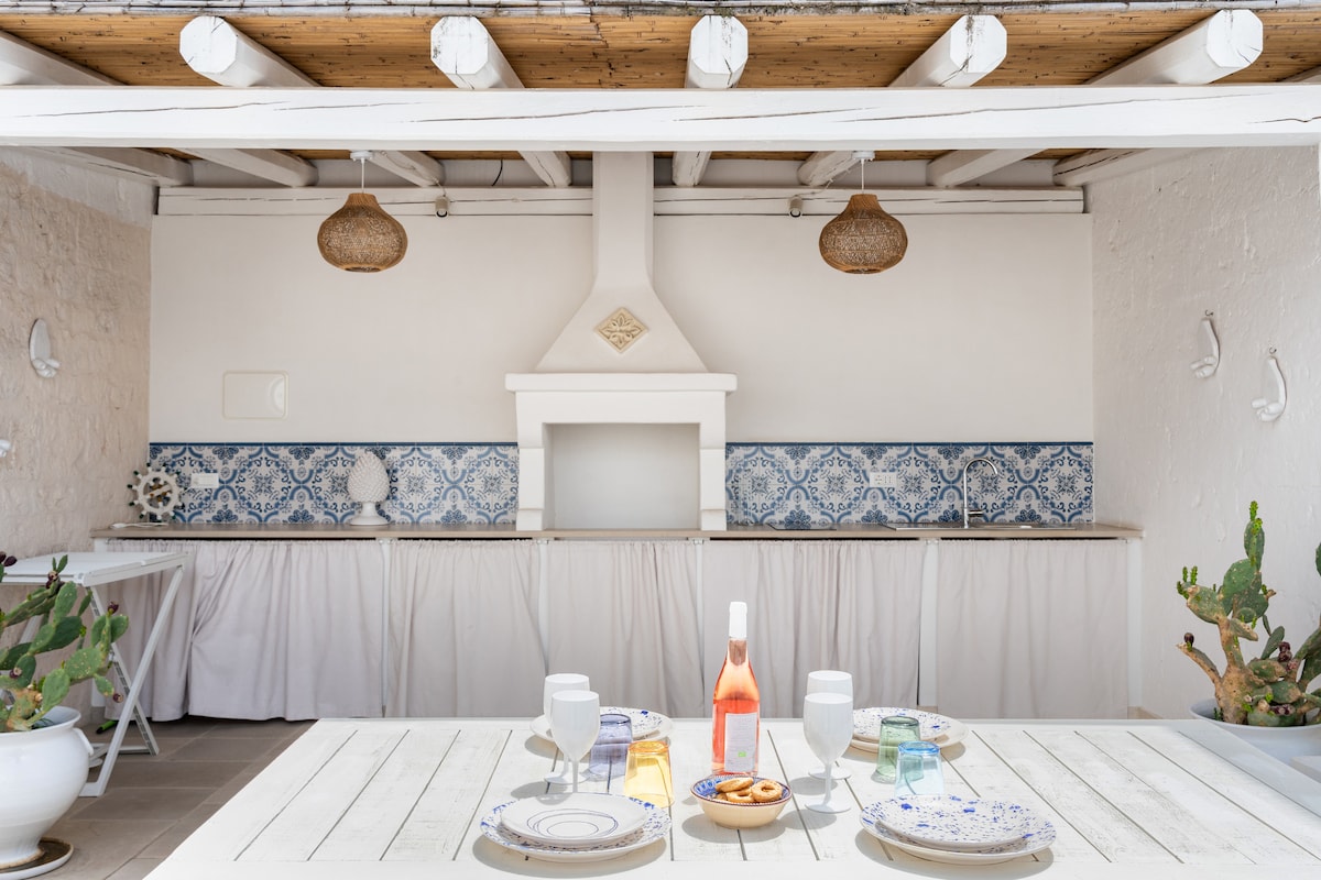 An outdoor kitchen area features a white stone oven and decorative blue tiles along the backsplash. A wooden table is set with plates, glasses, and a bottle, complemented by light-colored drapes. Natural light illuminates the space, enhancing the serene ambiance.