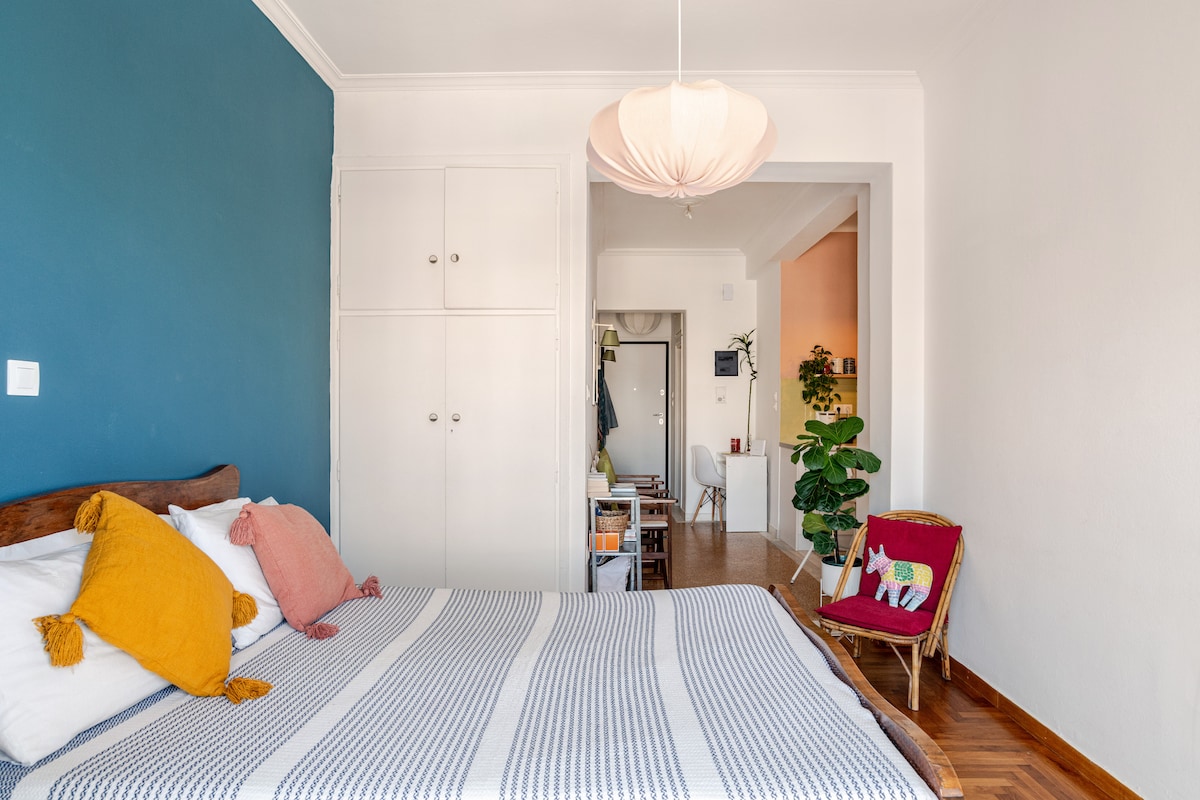 A cozy bedroom features a spacious bed adorned with striped bedding and colorful pillows. Light walls contrast with an accent wall in a calming blue. A simple light fixture hangs overhead, and a small entryway leads to an inviting living space visible in the background.