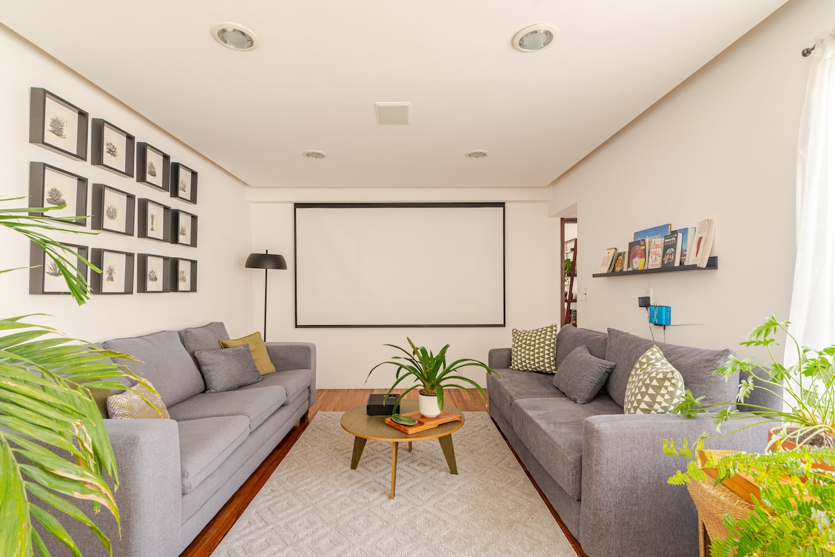 The living room presents two gray sofas arranged around a round coffee table, with a projector screen mounted on the wall. A collection of framed artwork adorns the wall, while plants add a touch of greenery to the space. Natural light filters through a nearby window.