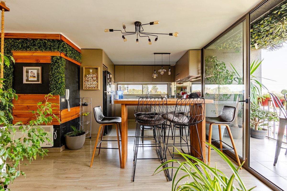 A modern kitchen and dining space features an island with bar stools, pendant lighting, and a spacious layout. Greenery is visible along the walls, adding a natural element. Large glass doors open to a terrace, enhancing the light-filled environment.