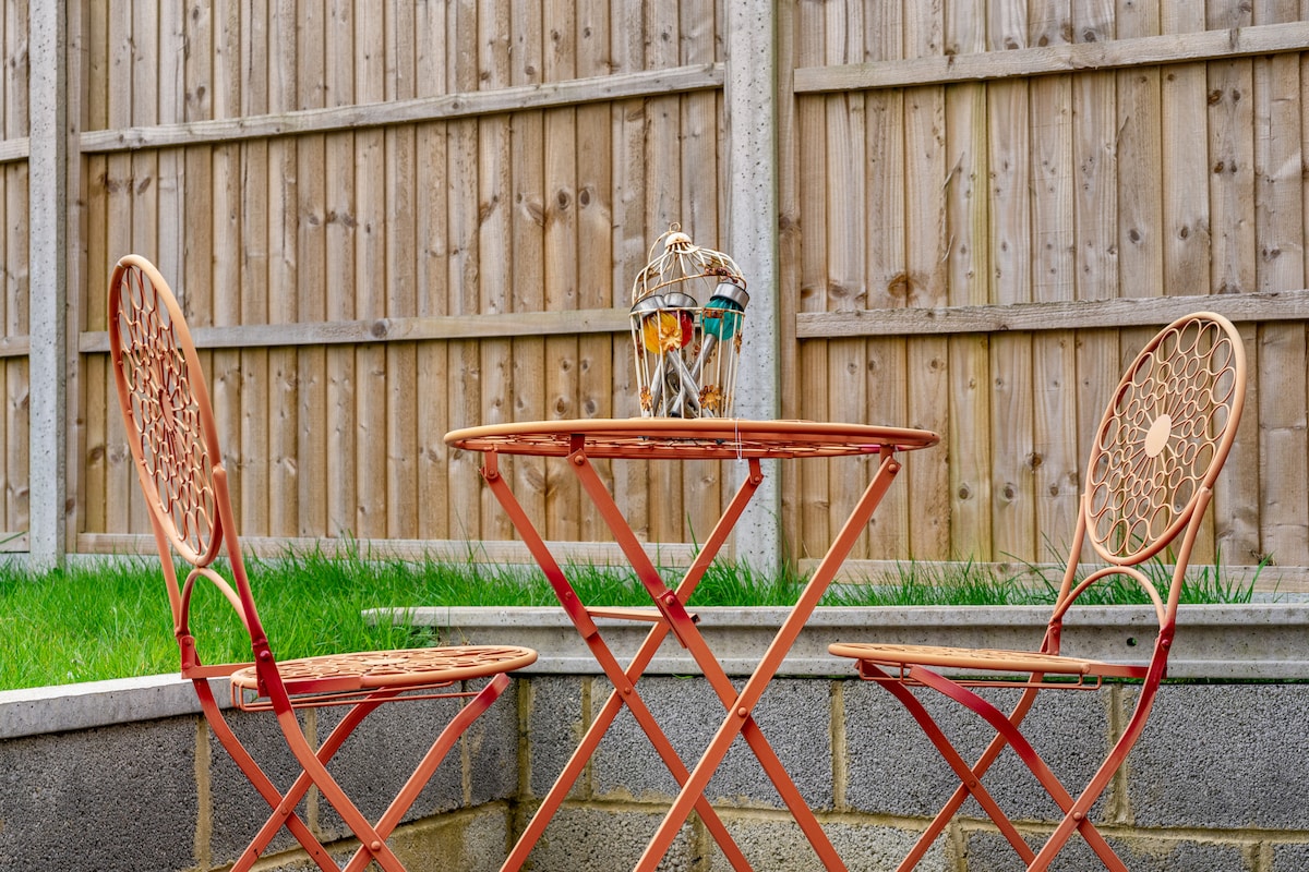 A compact outdoor seating area features a round table paired with two matching chairs, all in a warm reddish hue. The backdrop consists of a wooden fence, while soft green grass surrounds the seating arrangement, providing a serene space for relaxation.