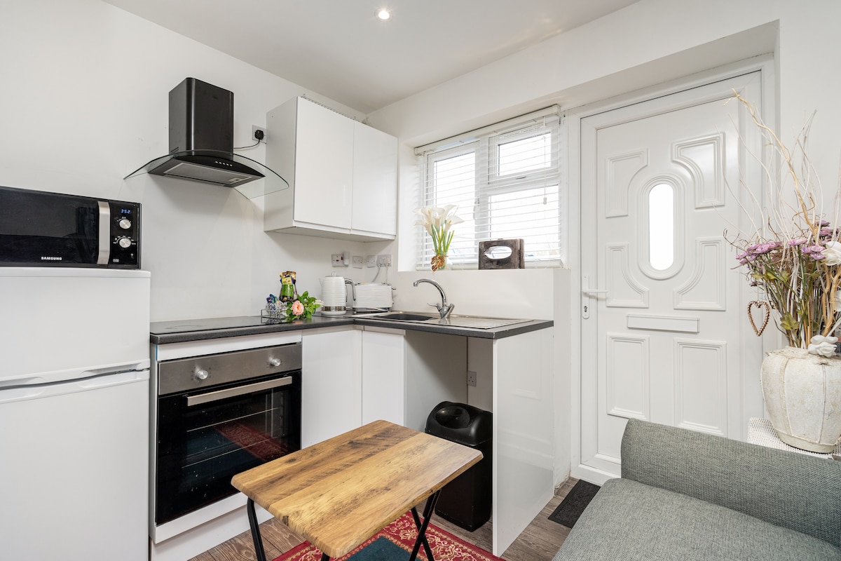 A compact kitchen area features modern appliances, including a microwave and an oven. A small wooden table is positioned in front of a gray sofa. Natural light enters through the window beside the sink, creating a functional space for meal preparation.