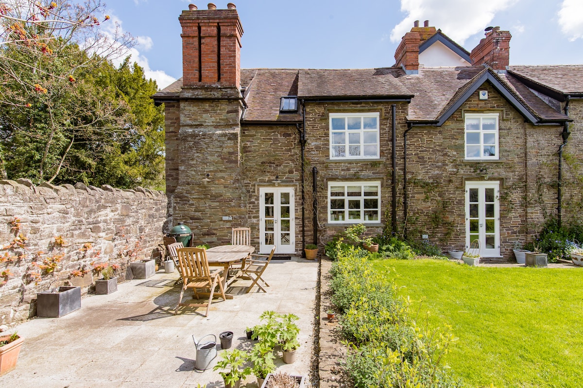 A charming exterior of the house is presented, featuring a walled garden with a spacious patio area. Wooden garden furniture is arranged around a table. The stone walls and multiple windows enhance the inviting atmosphere, all under a clear blue sky.