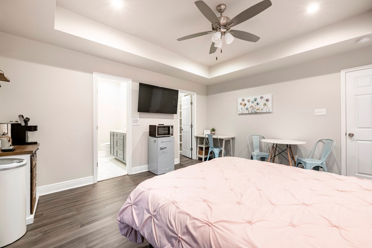 A spacious room is equipped with a large bed featuring a soft pink quilt. A ceiling fan is installed above, and a wall-mounted TV is visible. A small dining area includes two blue chairs around a round table. A private bathroom entrance is located in the background.