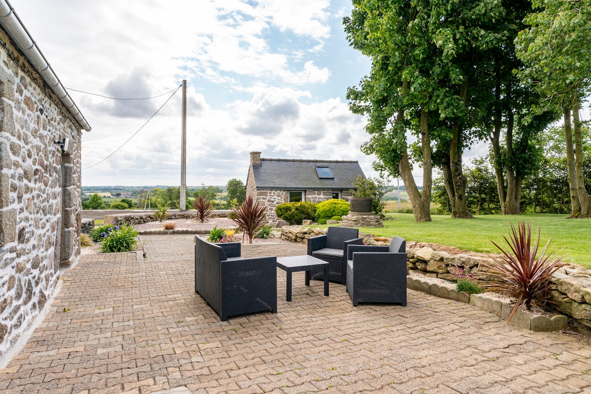 An outdoor patio area features a set of black wicker chairs and a matching table, positioned on a stone-paved surface. Lush greenery and well-kept landscaping surround the space, with trees and distant countryside visible under a partly cloudy sky.