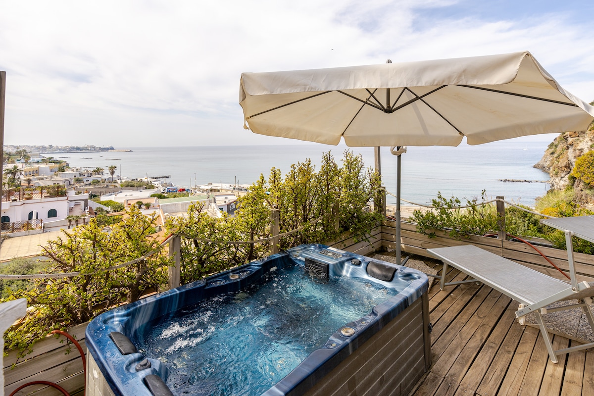 A private hot tub is featured on a sun-soaked terrace, surrounded by greenery. Sun loungers are positioned nearby, providing a comfortable spot for relaxation. The distant seascape offers a serene view of the coastline under a clear sky.
