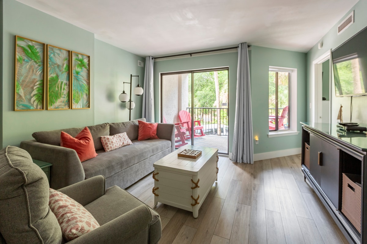 The open living area features a comfortable gray sofa and a cozy armchair, complemented by decorative pillows. A coffee table sits centrally, and natural light filters through sliding glass doors that lead to a private balcony. Tropical-themed artwork adorns the walls.