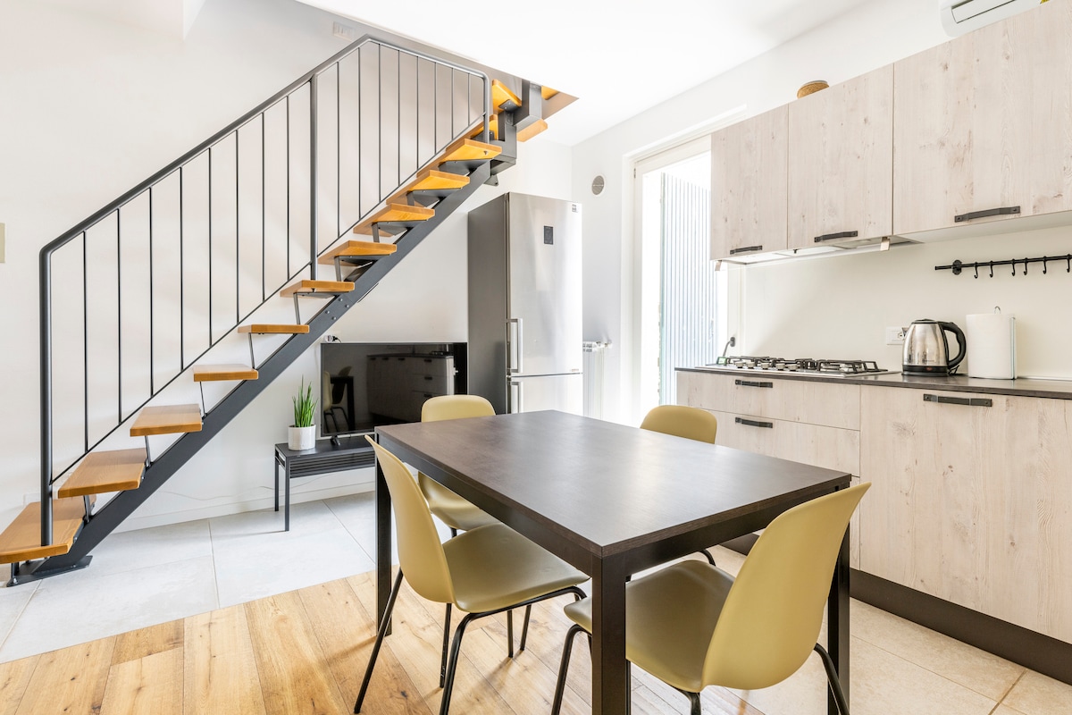A modern kitchen area is shown, featuring a dark wood table surrounded by four light-colored chairs. A staircase with wooden steps is visible in the background, leading to the upper floor. A refrigerator and a smart TV are seen along one wall, complementing the open layout.