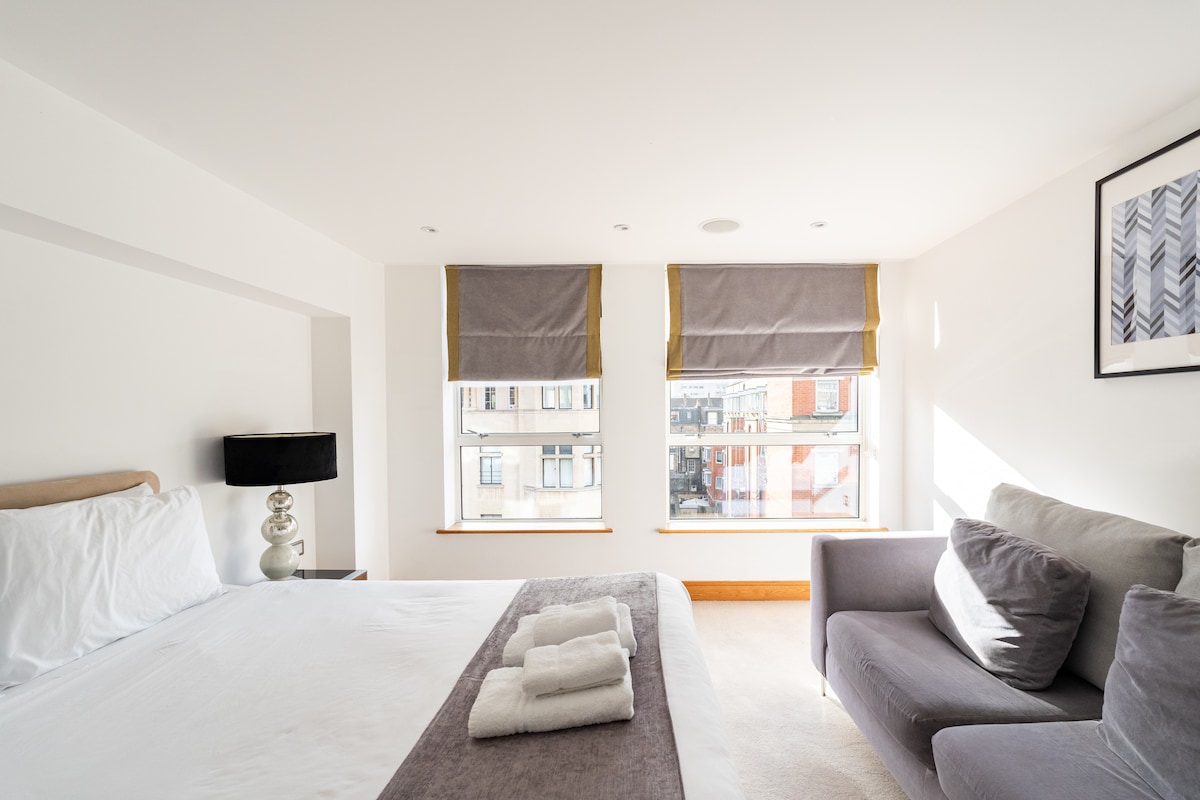 The main bedroom features a spacious king-size bed dressed in white linens, accompanied by soft towels neatly arranged. Two windows allow natural light to fill the room, with light grey window treatments framing the views. A comfortable grey sofa offers additional seating beside the bed.