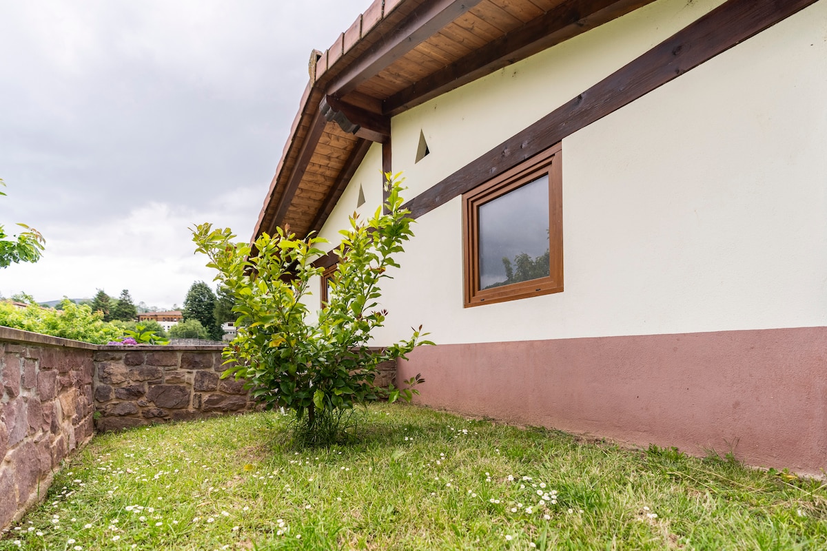 A corner of the exterior showcases a stone wall leading up to a wooden structure with a sloped roof. A small tree is positioned in the grassy area, framed by fresh greenery and scattered flowers, while a window offers a glimpse of the interior.