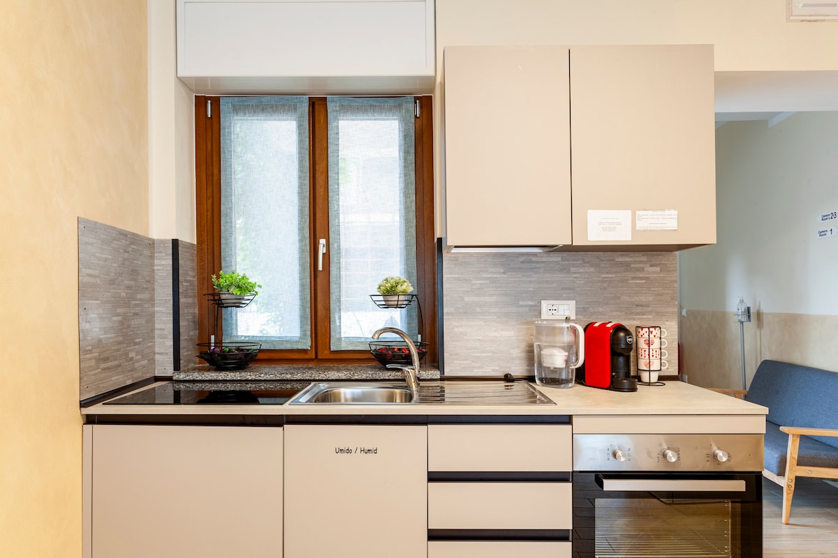 The kitchen area features a minimalist design with light cabinetry and a sleek countertop. A window above the sink allows natural light in, while essential appliances, including a coffee maker and oven, are positioned conveniently. Decorative plants enhance the space.