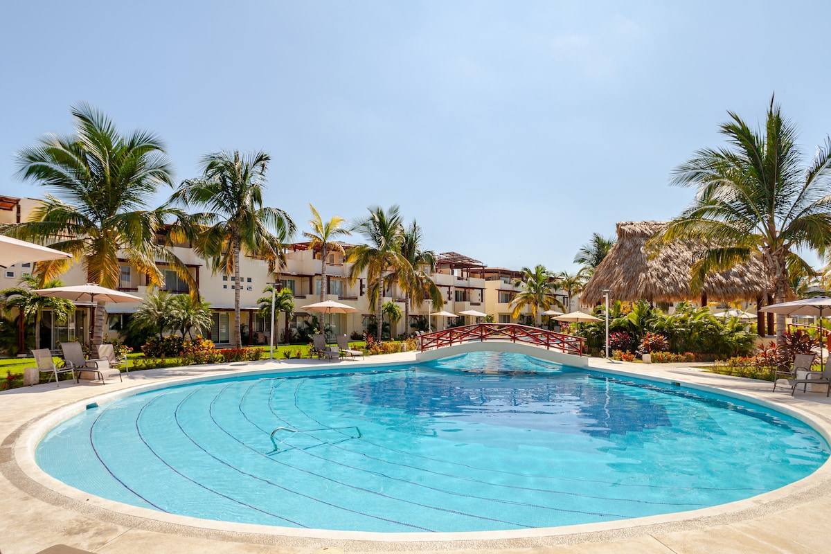 A curved pool surrounded by lush landscaping features palm trees and tropical plants. Comfortable lounge chairs are placed under umbrellas, and a thatched palapa offers shaded seating. Nearby, a small wooden bridge crosses the pool, connecting paths through the serene outdoor area.