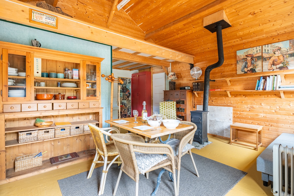 A cozy dining area features a wooden table set for four, complemented by woven chairs. A wooden sideboard displays dishes and storage baskets. Decorative art hangs on the wooden walls, enhancing the rustic ambiance. A small wood stove is positioned in the corner, providing warmth.