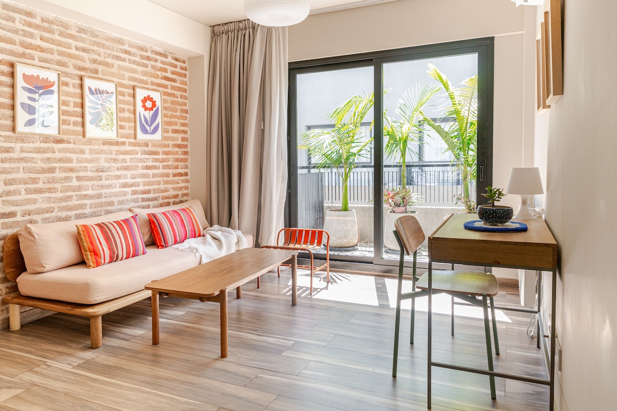 A light-filled living area features a comfortable sofa adorned with colorful striped cushions. A wooden coffee table sits in front of it, and a desk is positioned nearby. Large glass doors open to a small outdoor space with greenery.