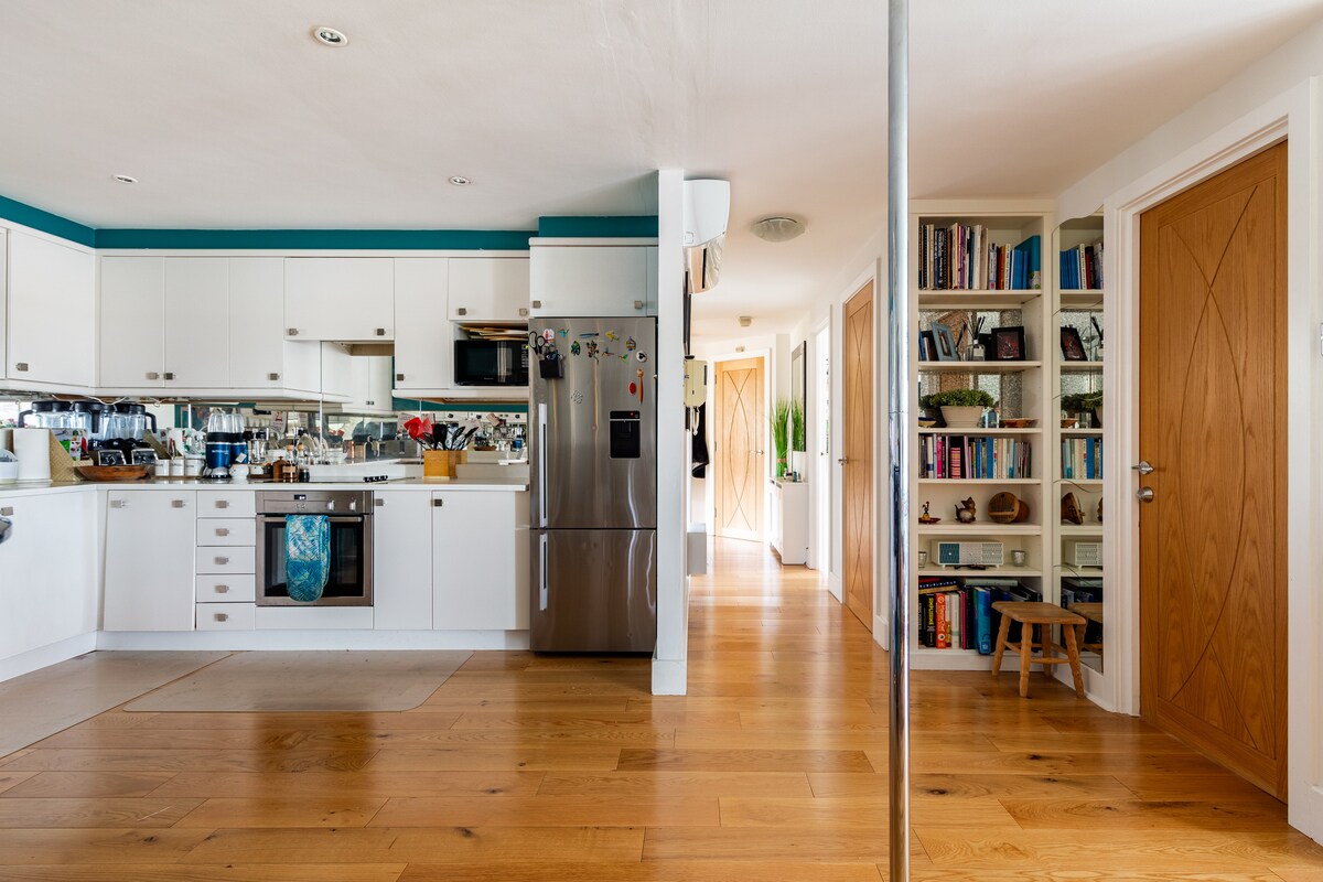 A spacious kitchen area with modern appliances, including a stainless steel refrigerator and oven. White cabinets are complemented by wooden flooring, creating a warm ambiance. A bookshelf filled with books is visible in the hallway, leading towards the entrance doors.
