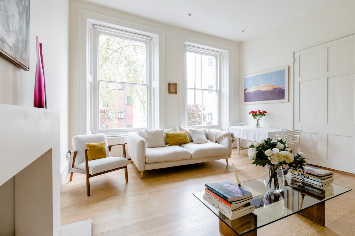 A bright living area features large windows allowing ample natural light. A light-colored sofa is complemented by cushions. A contemporary coffee table displays books. A dining table in the background is set for four with a vase of flowers, and light wooden floors add to the airy atmosphere.