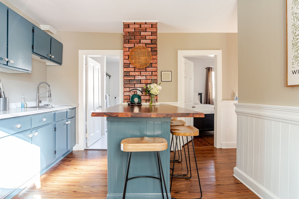 The kitchen features blue cabinetry with a wooden countertop extending into a breakfast bar with three stools. A brick accent wall adds texture, while natural light enhances the inviting atmosphere. Doorways lead to adjoining areas, and fresh flowers sit on the countertop.