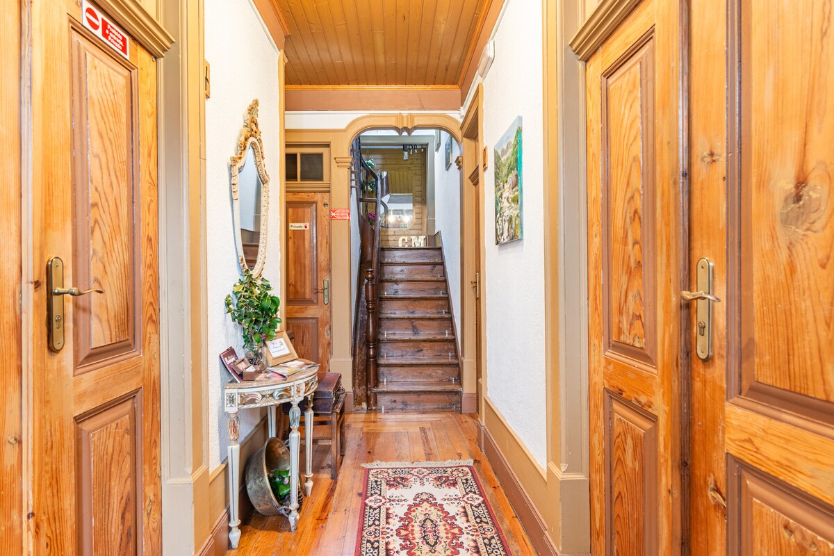 A well-lit hallway features wooden doors and a staircase leading to the upper levels. A decorative mirror and potted plant are positioned beside a small table. A patterned rug enhances the entryway's warmth, while wooden paneling and detailed moldings contribute to the inviting atmosphere.