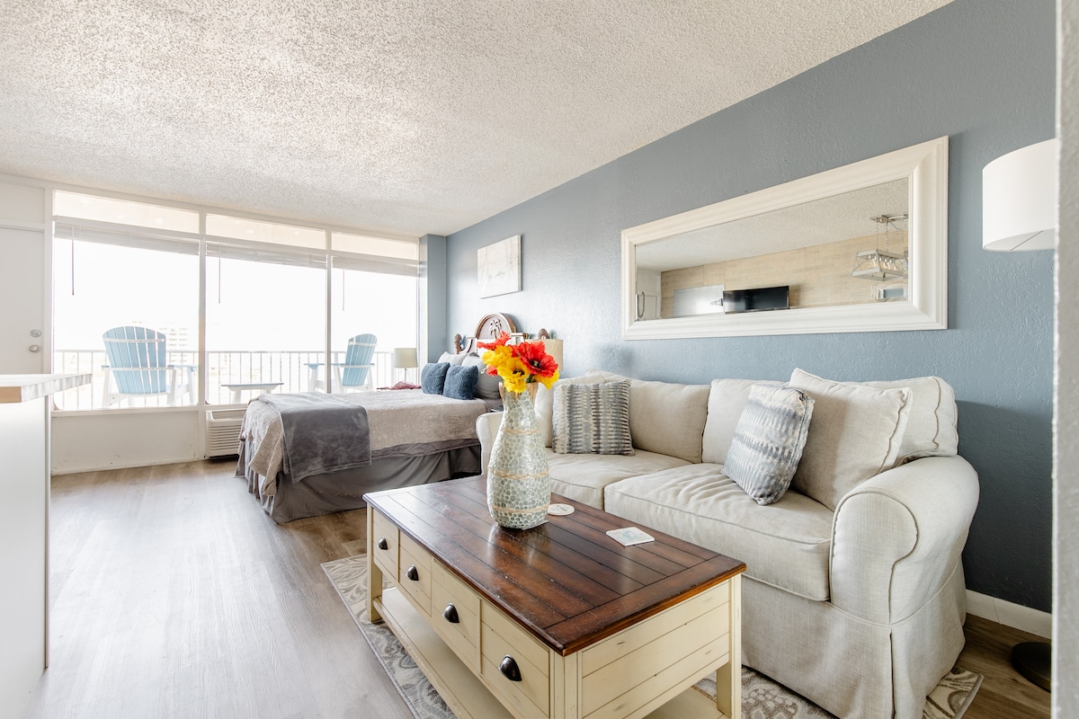 A bright and airy studio space features a light-colored sectional sofa and a wooden coffee table adorned with a vase of colorful flowers. Large windows offer views of the ocean and fill the room with natural light, complemented by soft, neutral walls and bedding.