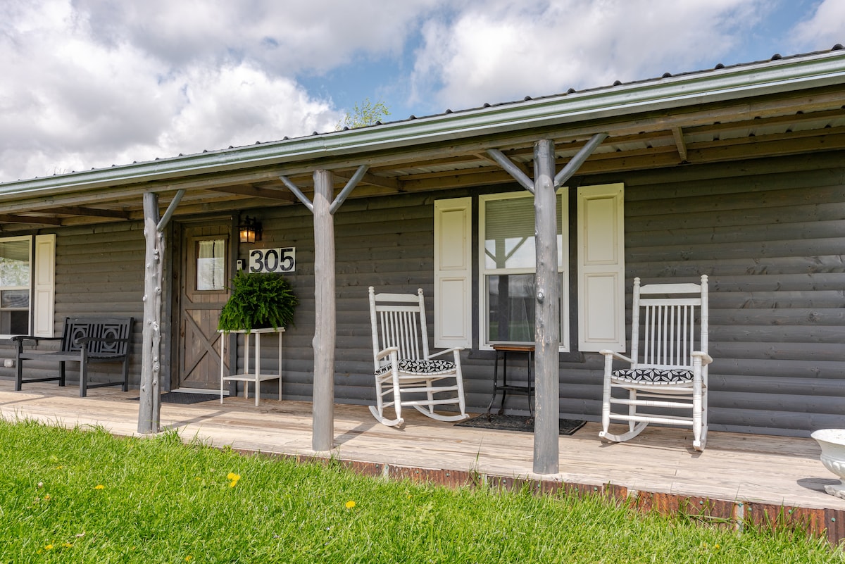 The front porch features two classic rocking chairs positioned beside a small table, surrounded by well-maintained grass. Light-colored shutters frame the windows, and a welcoming cabin number is displayed prominently. The cabin's log siding contributes to its rustic charm.