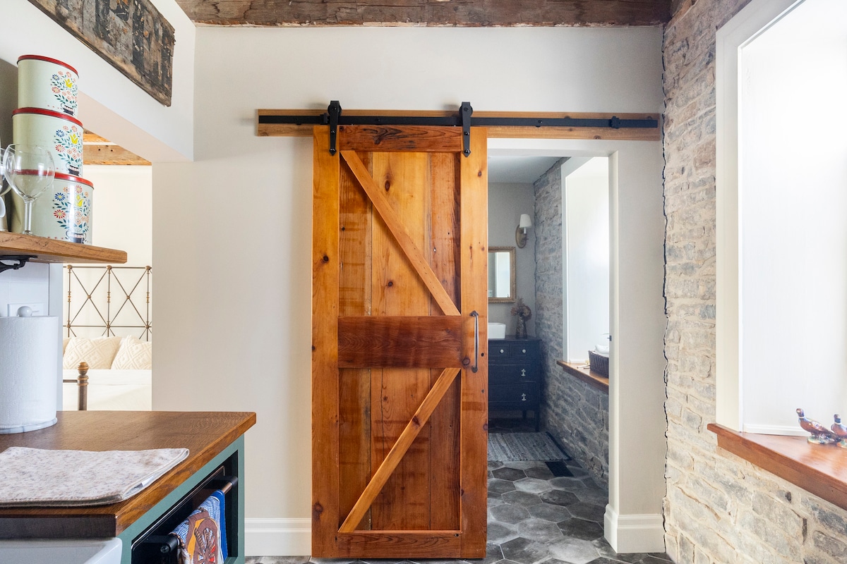 A rustic wooden sliding barn door, featuring a rich brown finish, is positioned in an open space with exposed stone walls. Natural light fills the area, highlighting the texture of the stone and the warmth of the wooden beams above.