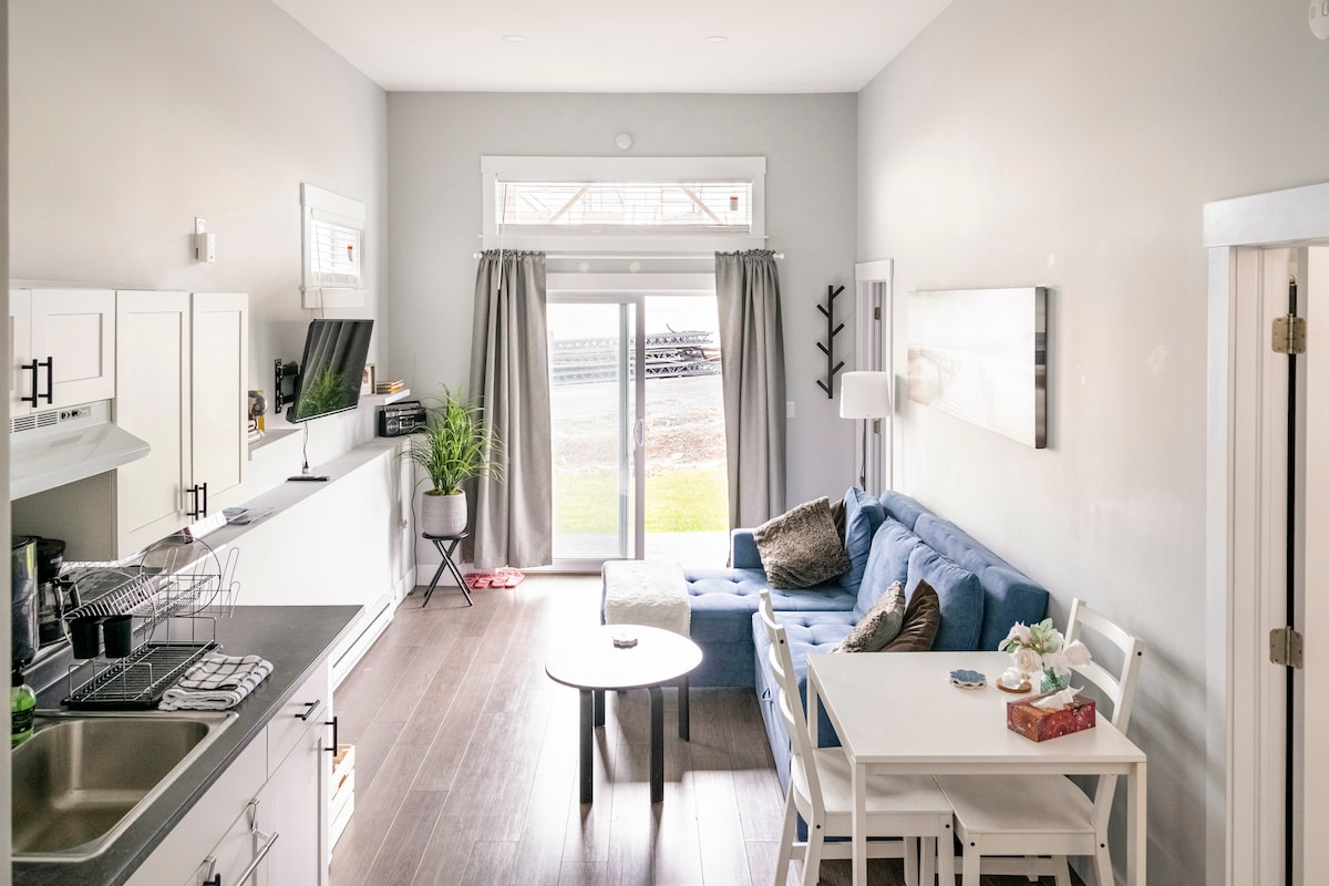 A bright living space features large windows allowing natural light to flood the room. A sectional sofa and small dining table provide comfortable seating. The kitchenette is equipped with essential appliances, and light grey walls create a neutral backdrop for the furnishings.